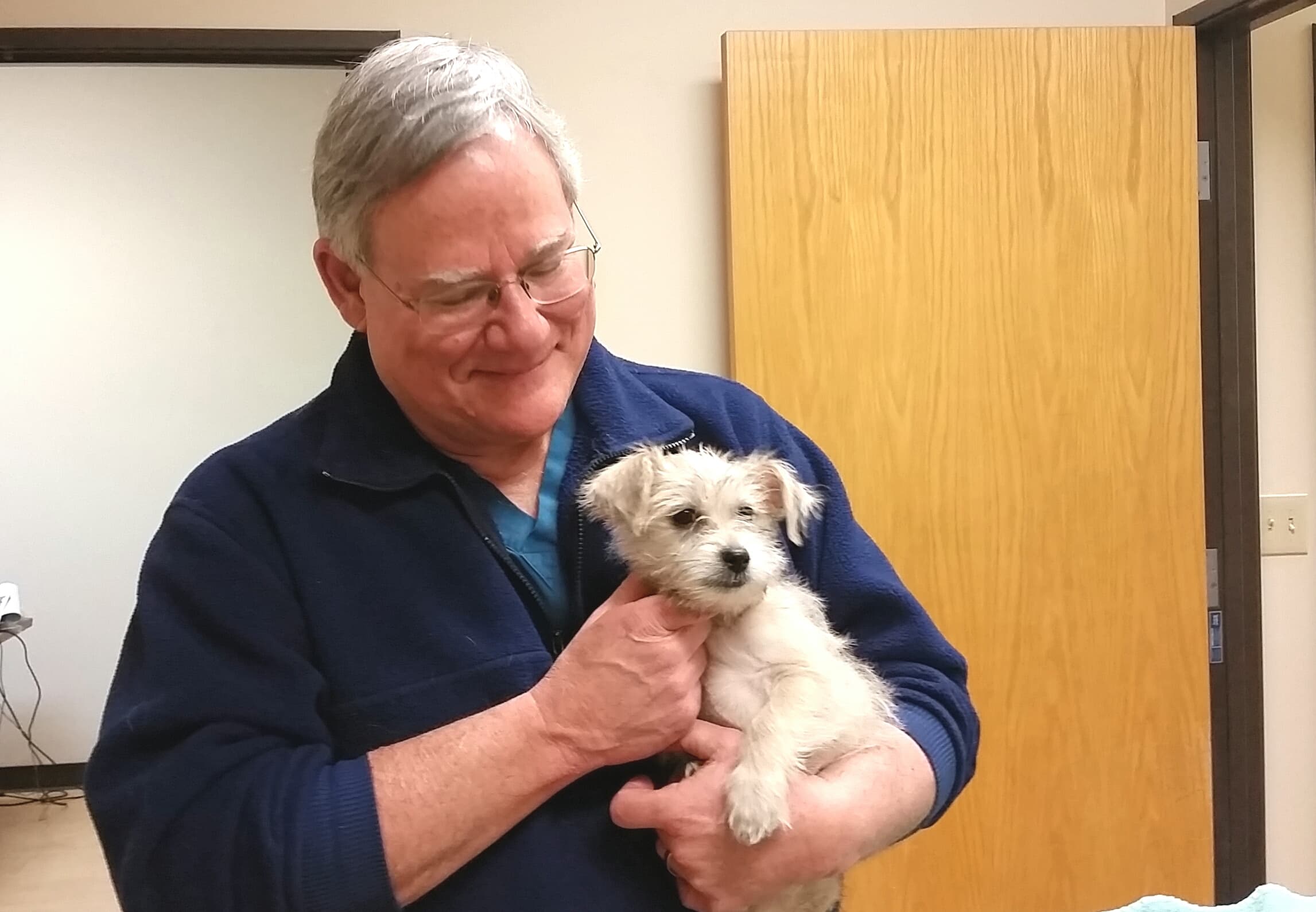 Dr. John Summar smiling down at a small white terrier puppy he is holding in an exam room