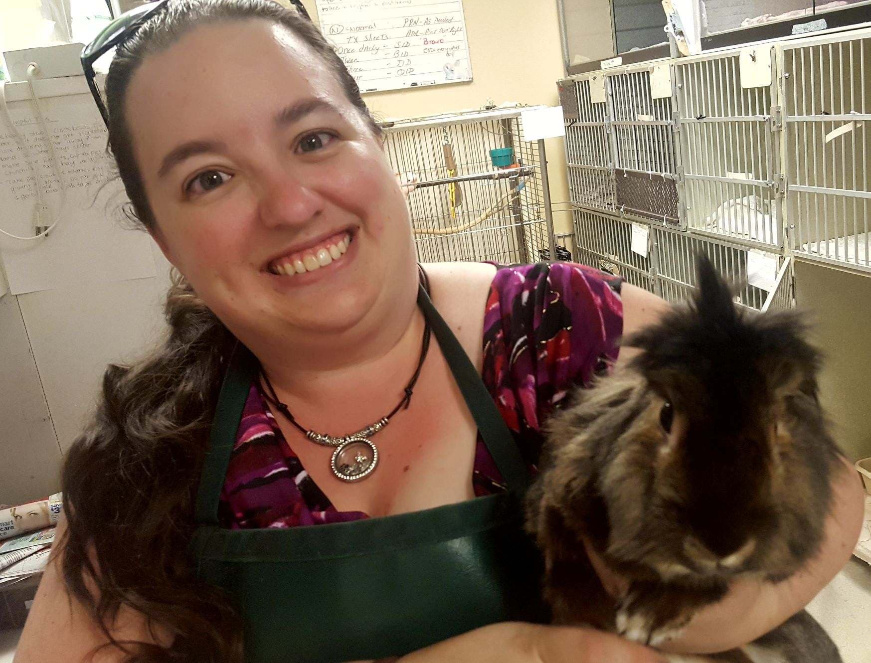 A smiling staff member in a green apron holding a long-haired guinea pig, with animal cages behind her